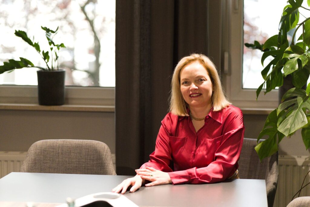Olga Müller, Gründerin von Womenmatic, sitzt lächelnd an einem Besprechungstisch, im Hintergrund Fenster, Vorhang und Pflanzen